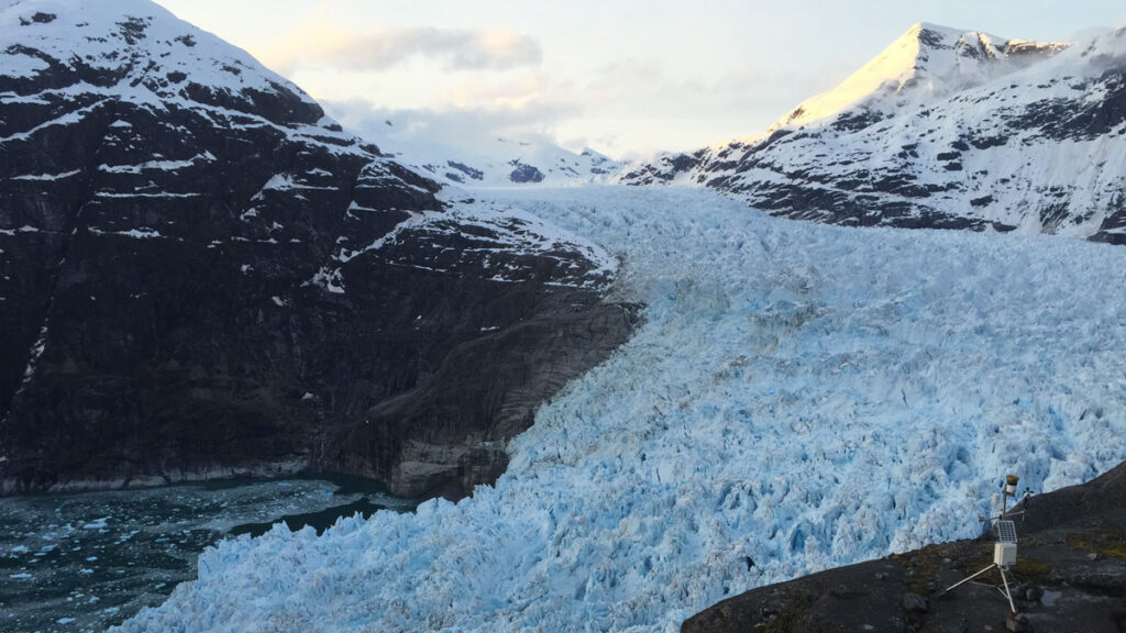 A weather station stands atop a rock outcrop in the foreground, above a glacier flowing between mountains and into the water.