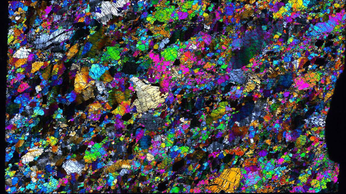 Image of a thin section of peridotite, taken under a microscope, with the pinks, greens, purples, and blues of olivine crystals of various sizes mixed with other, less brightly colored minerals