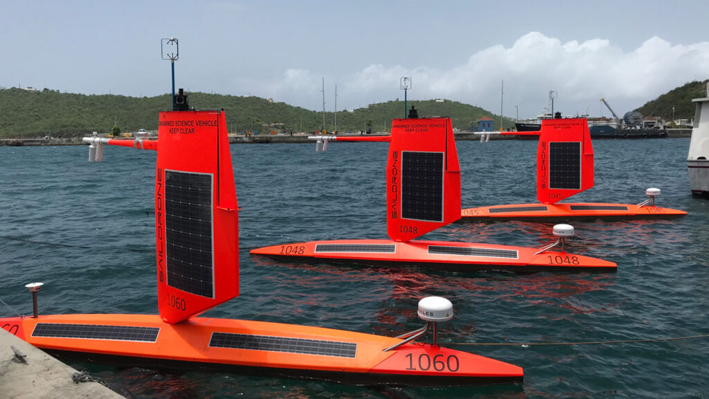 Three red saildrones with built-in solar panels float in a line in the water beside a dock.