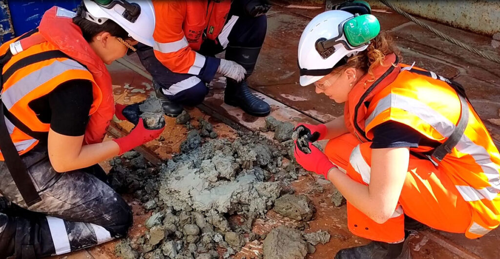 Researchers crouching down and looking closely at samples of rock and sediment on the deck of ship