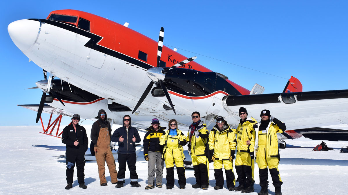 A team of researchers stands in front of an airplane in Antarctica.