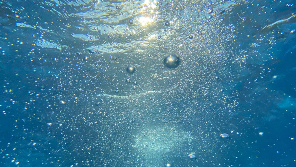 A view from underwater, looking through blue water, ripples, and bubbles toward light at the surface