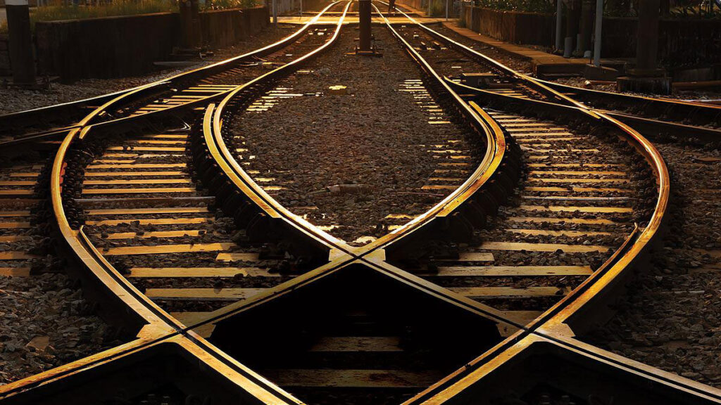 Two train tracks cross and then bend to run parallel to each other at sunset.