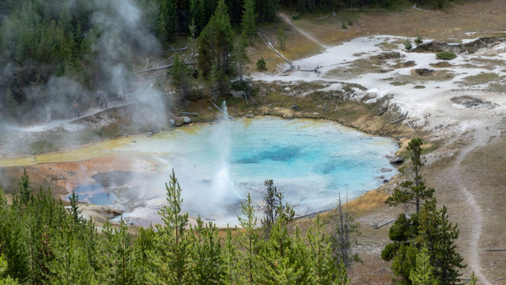 Una imagen del géiser Imperial de Yellowstone. El géiser está al centro de la imagen mostrando coloraciones azules claras y en la parte inferior de la imagen se observan las copas de unos pinos.