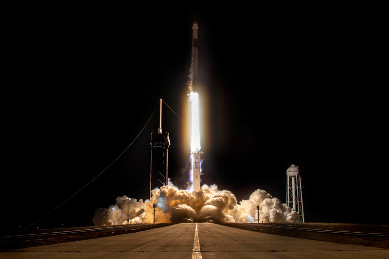 SpaceX Crew Dragon spacecraft launches vertically into space against a black night sky, with the bright lights of rocket fuel below, and billowing clouds on the ground. A road with a single pained line in the foreground leads directly to the launch.