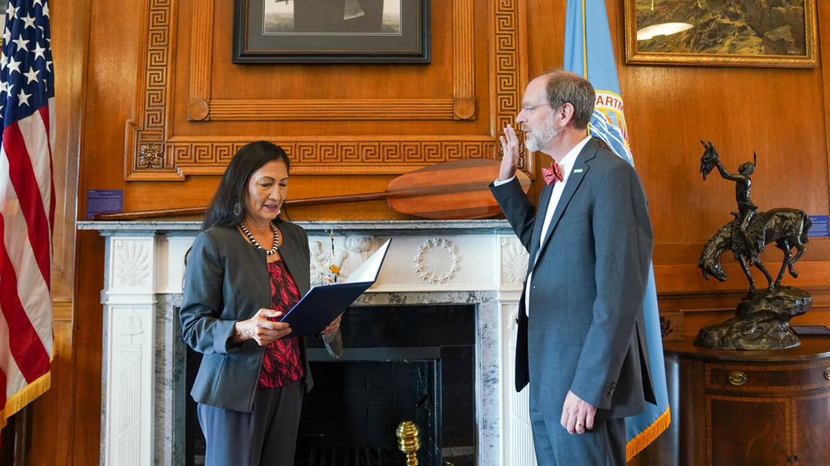 Deb Haaland, wearing a gray suit with a red and black blouse, reads from a black book as she swears in the new USGS director. David Applegate, wearing a gray suit, white shirt, and red bow tie, stands opposite her with his right hand raised as he is sworn in. The pair stand in front of a large white marble fireplace set in a wood-paneled wall. The wall is decorated with Native American artwork, and the fireplace is framed by an American flag and a light blue flag for the U.S. Department of the Interior.