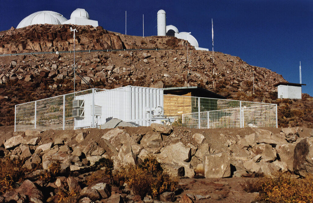 Several telescope domes and small shacks atop a rocky desert mountain.