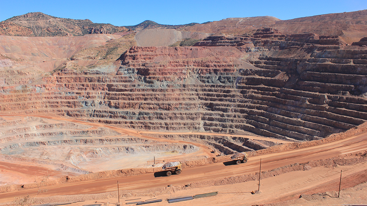 A copper mine delves deep into the orange rock of an Arizona desert. More than a dozen even steps have been excavated leading down to a roughly circular depression in the ground. Some of the steps have patches of oxidized copper, a blue-green color. A smooth path large enough for heavy machinery spirals down into the bottom of the mine.