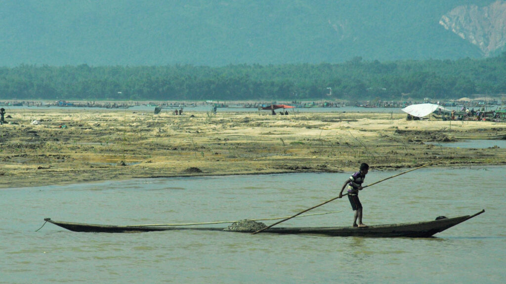 A boy in a striped t-shirt and shorts steers a long, narrow, and shallow boat with a long pole across a shallow body of water. The boat is laden with rocks. In the background is a marshland with a small group of people and a few tents, and beyond that are forests and mountains. One patch of forest on the right side of the mountains has been stripped bare by a past landslide.