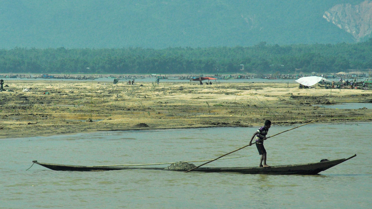 A boy in a striped t-shirt and shorts steers a long, narrow, and shallow boat with a long pole across a shallow body of water. The boat is laden with rocks. In the background is a marshland with a small group of people and a few tents, and beyond that are forests and mountains. One patch of forest on the right side of the mountains has been stripped bare by a past landslide.