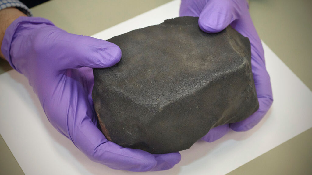 Gloved hands hold a dark chunk of rock, a part of the Aguas Zarcas meteorite.