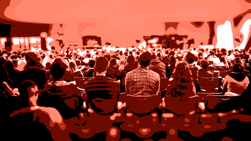 Image of many individuals seated at a conference. The photo was taken from the back of the room and shows the backs of the conference goers; it has a red, posterized treatment applied to it.