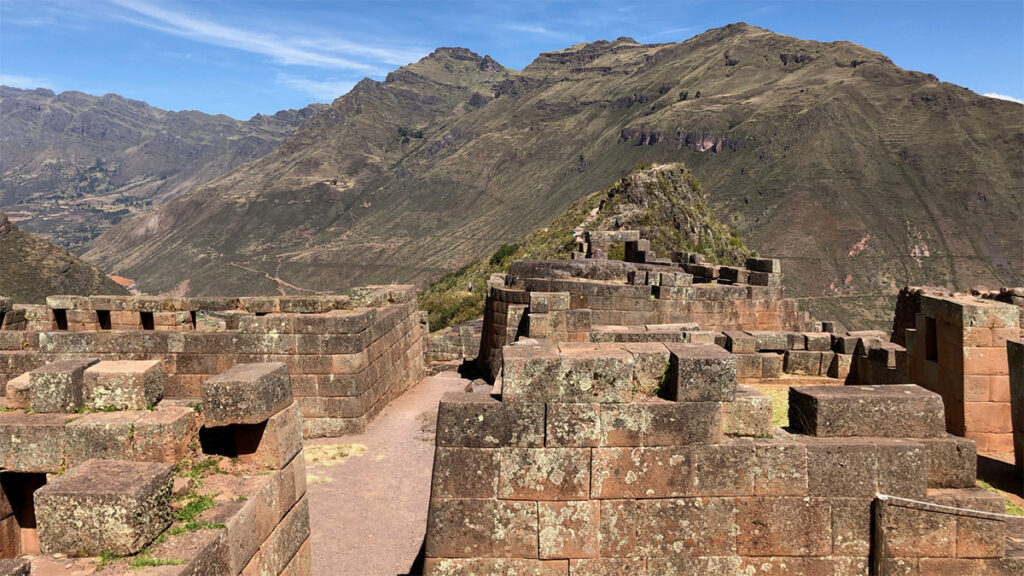 Photo showing Inca buildings in the foreground and mountains in the background; some cracks are visible in the stonework.