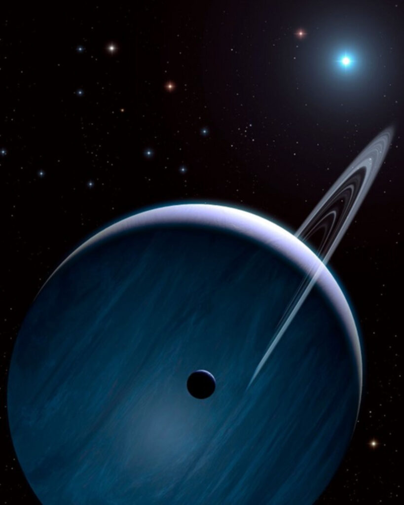 A gas giant planet in shades of blue and gray orbits a distant bright blue star. The planet has a ring system like Saturn’s that is seen almost edge on and is tilted diagonally from the top right toward the bottom left. The silhouette of one moon is visible in the foreground of the image. More stars in shades of blue, red, yellow, and white are strewn across the background.