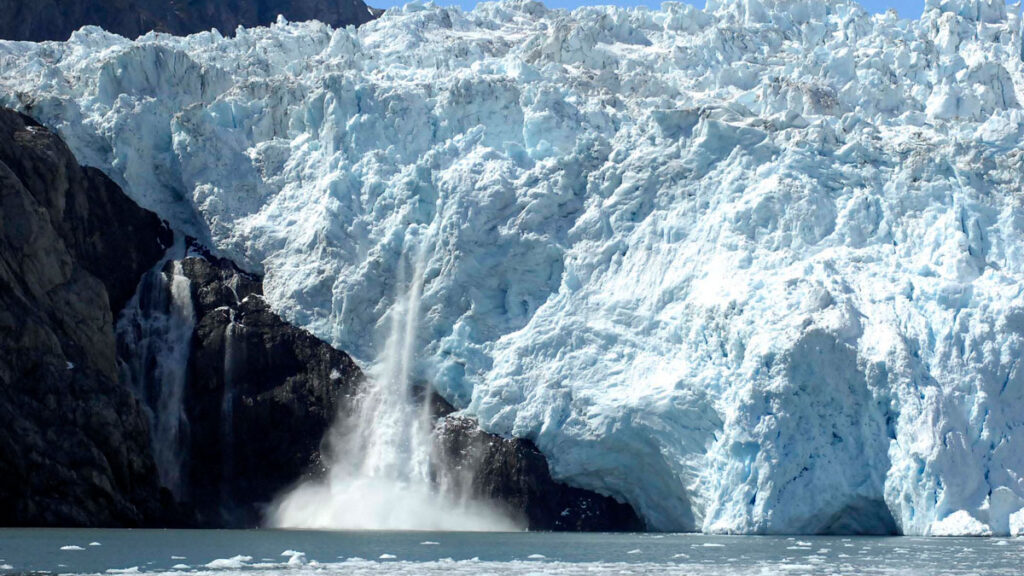 A large wall of ice looms above a body of water, and a waterfall trickles down the ice near the center of the image.