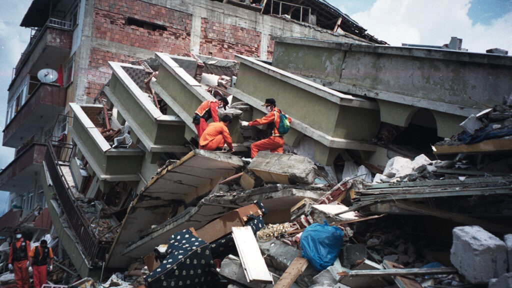 Tres rescatistas, vestidos en color naranja, buscan entre los escombros de un edificio caído. Detrás del edificio caído hay otro edificio de ladrillos rojos, el cual aún se encuentra de pie, pero tiene algunas paredes dañadas.