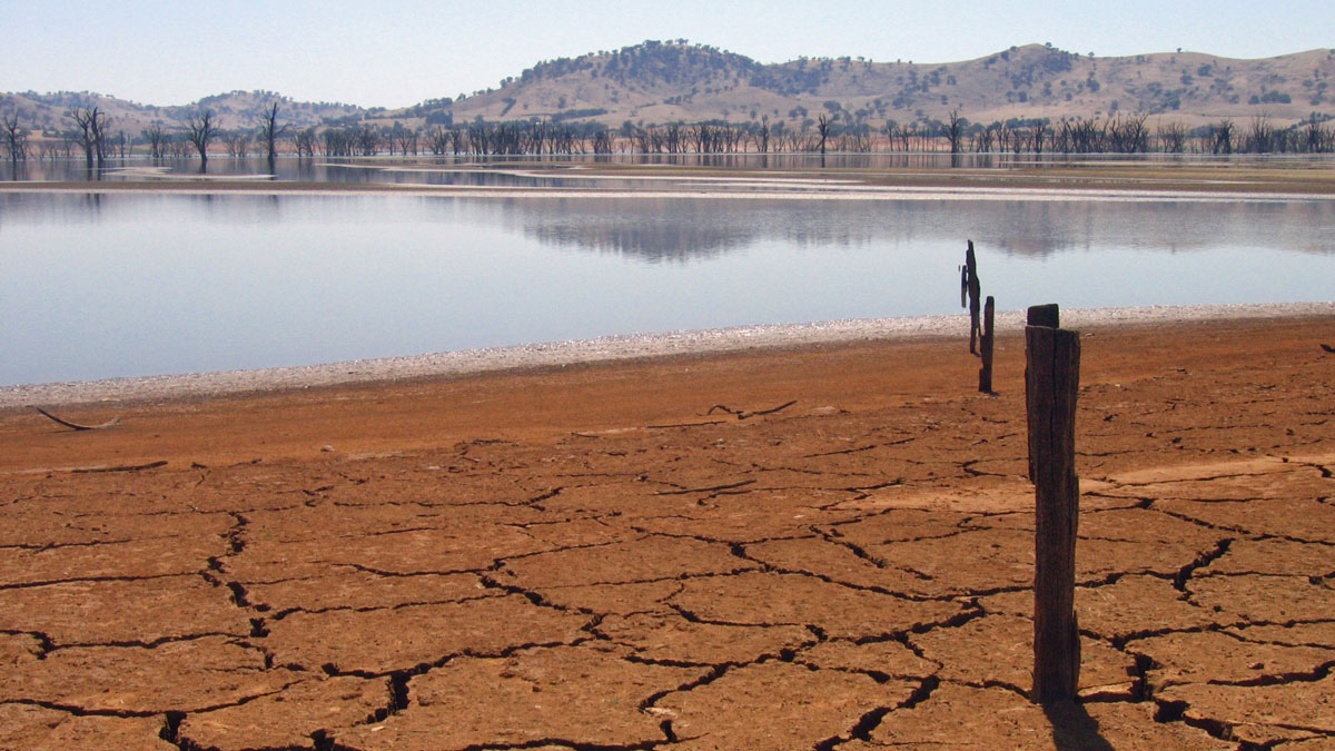 View of a lake in the distance with mud cracks in reddish soil in the foreground.