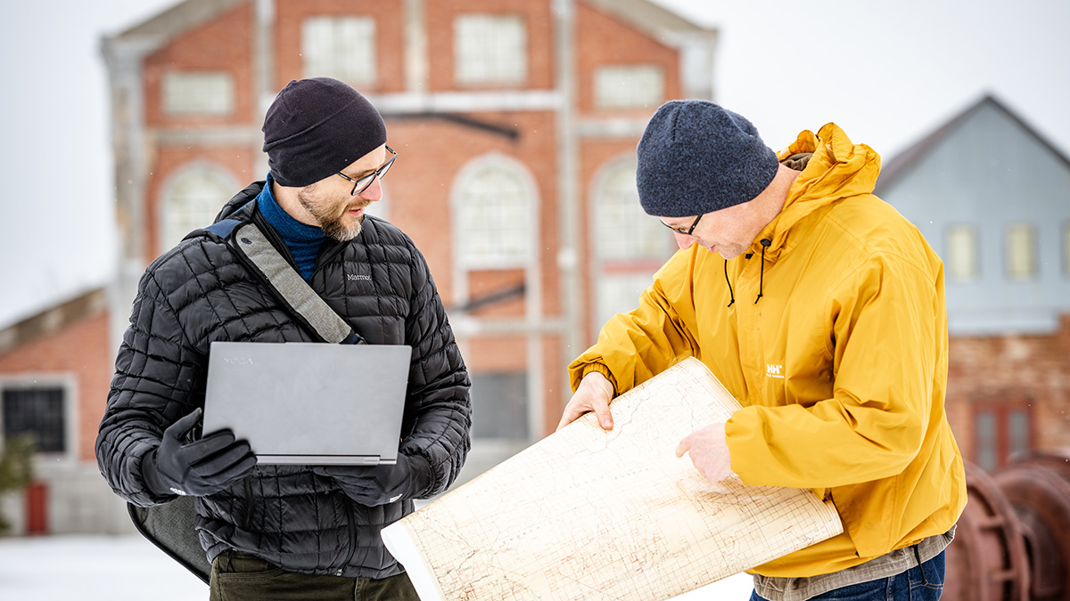 Researchers Roman Sidortsov and Timothy Scarlett stand in the snowy weather looking at a historical map of a mine site.