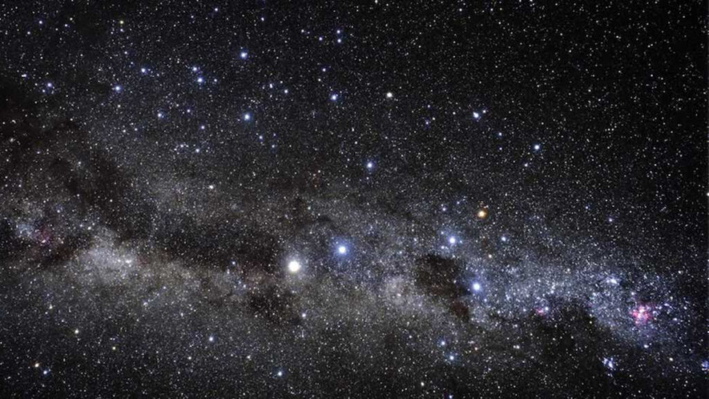 An optical astronomy image shows hundreds of stars in shades of blue, white, yellow, and red, with a dark band of dust running horizontally across the image. The stars are a range of sizes, from bright blue spots to no more than pinpricks.