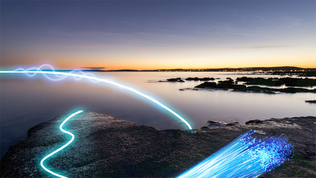 Image overlooking a rocky coastline and water just after sunset with a bright blue line representing a fiber-optic cable curving over the water and rocks