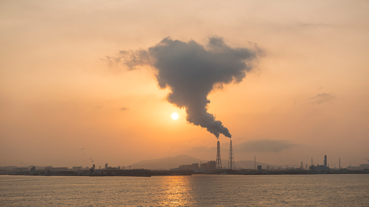 Emissions rise from industrial towers along a waterfront in front of a hazy orange sunset.