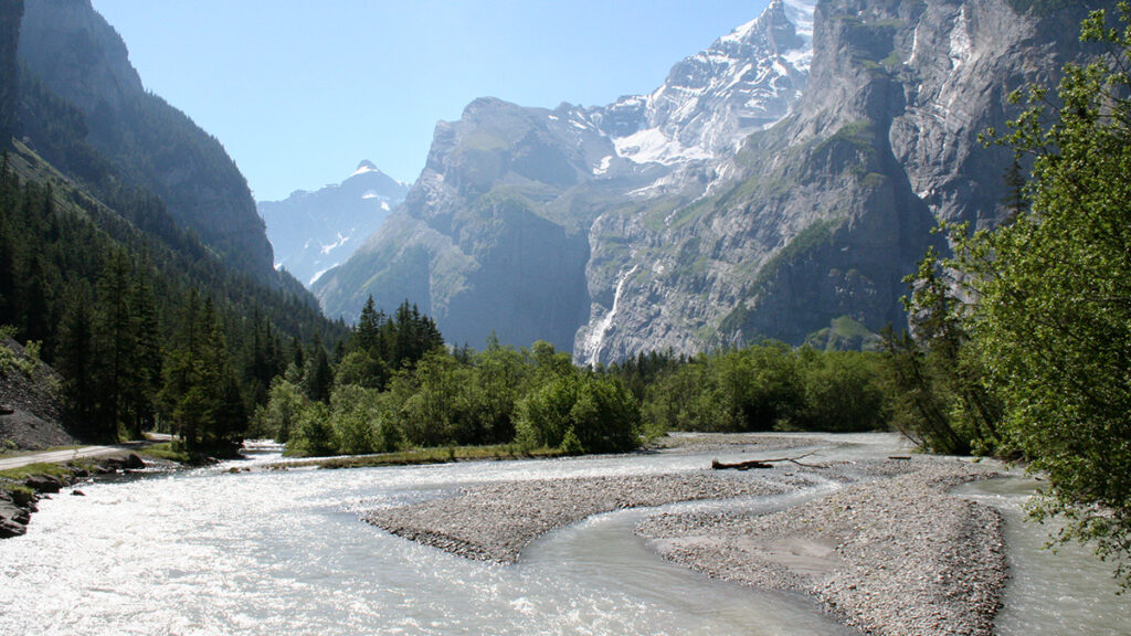 A blue-gray river runs through a steep mountainous terrane, with the slopes nearest the river coated in green grasses, plants, and coniferous trees.