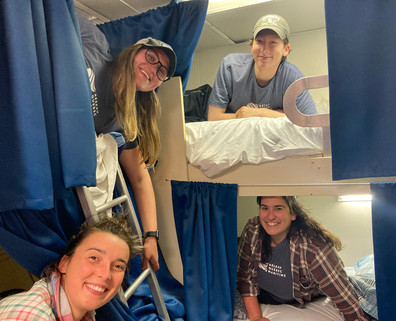 Four people smile at the camera from ship’s bunks.