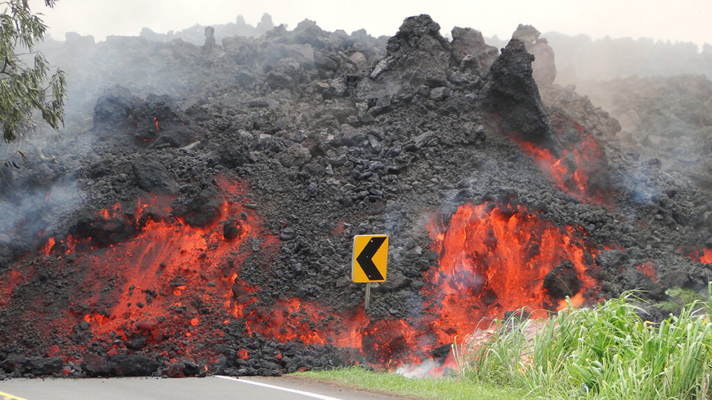 A mass of steaming, orange-glowing lava consumes a street sign as it flows over a roadway.