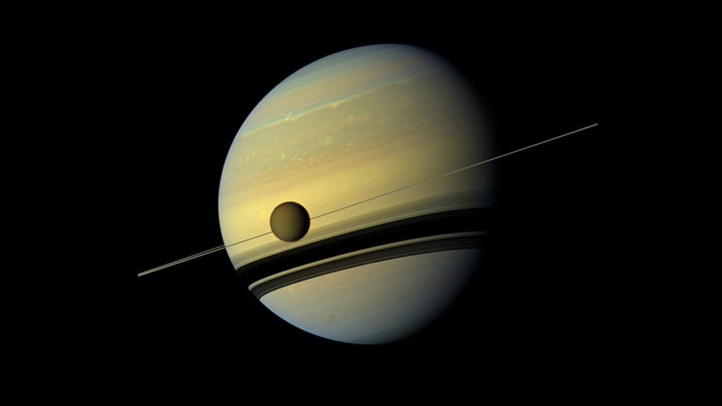 The orange moon Titan passes in front of the striped disk of the planet Saturn, with Saturn’s rings seen edge on, forming a thin line between the moon and planet.
