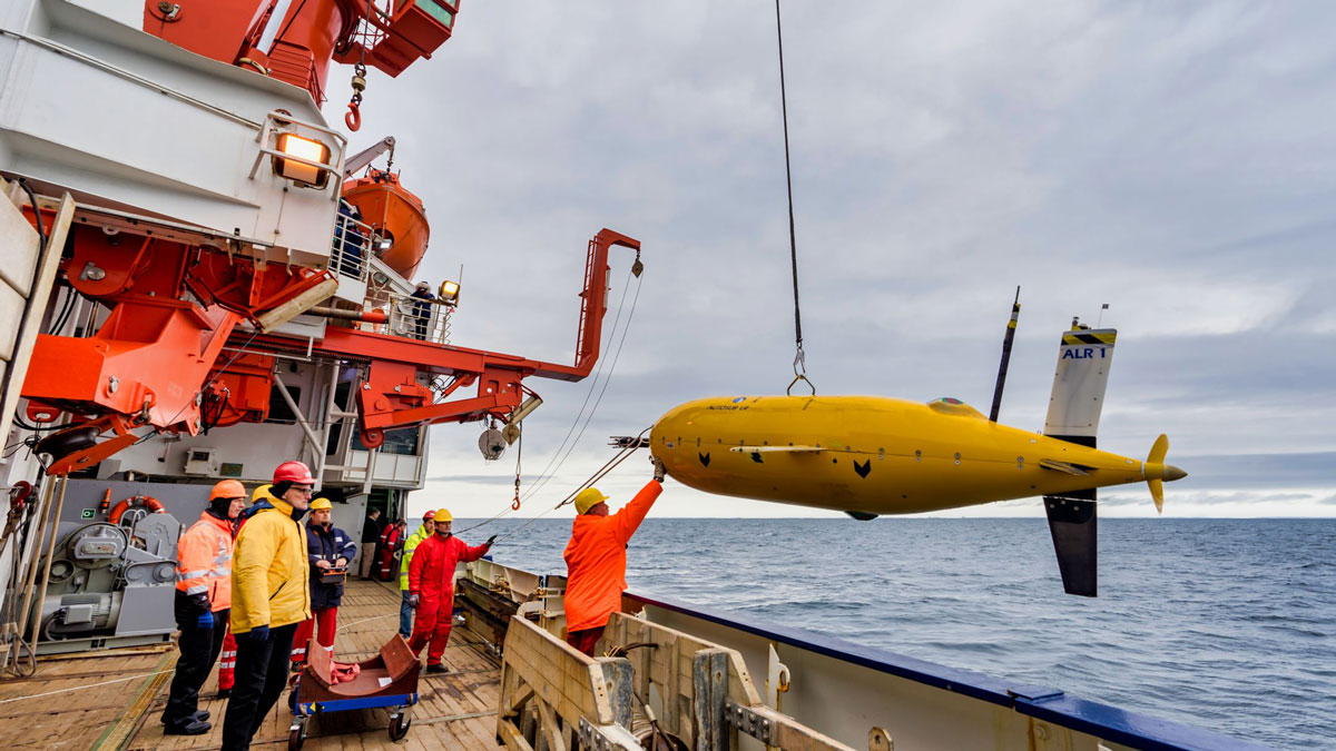 A yellow submarine-shaped autonomous underwater vehicle hangs from a cable over the side of ship as technicians and scientists look on.