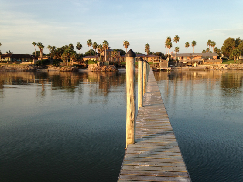 A long dock stretches over a water body with large residential houses and palm trees in the background.