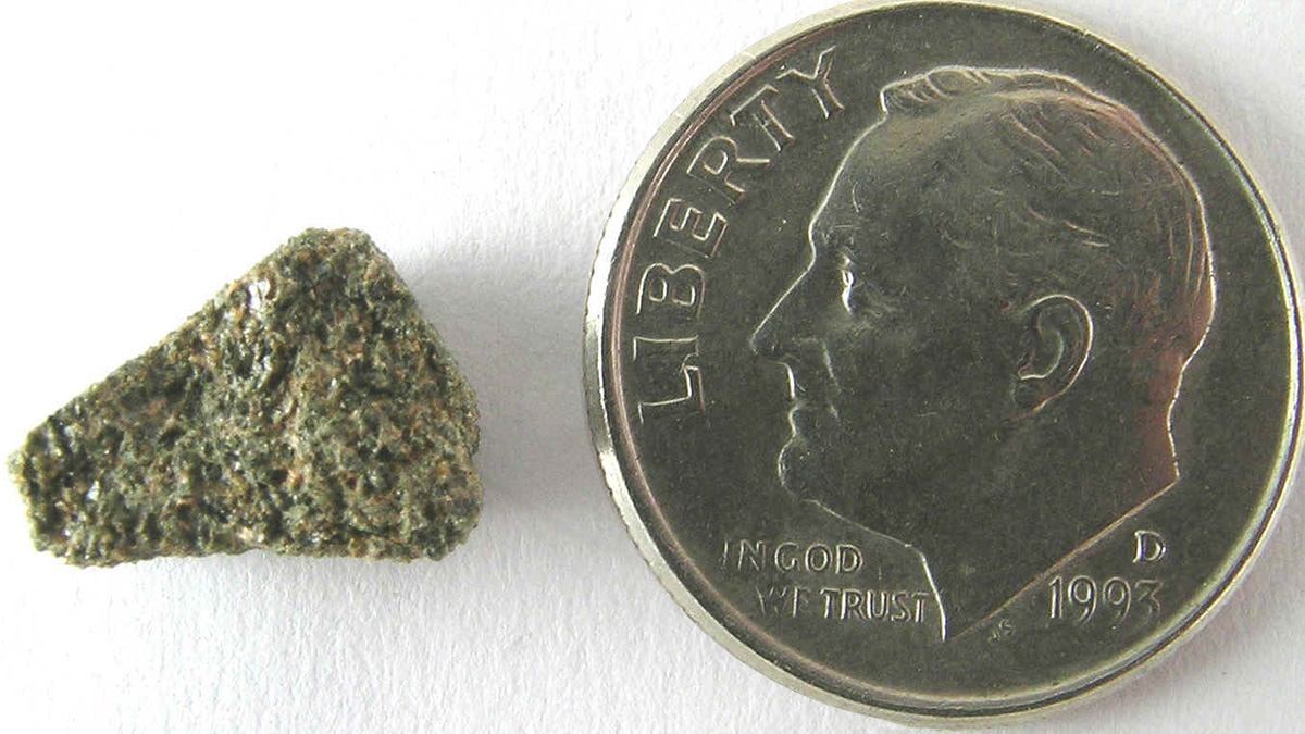 A pebble-sized meteorite sits next to a dime for scale.