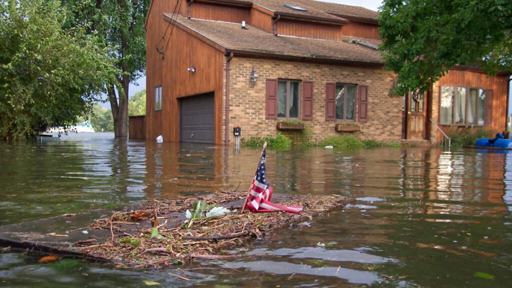 View of a house surrounded by floodwaters, with a piece of wood topped by a small United States flag floating in the foreground.