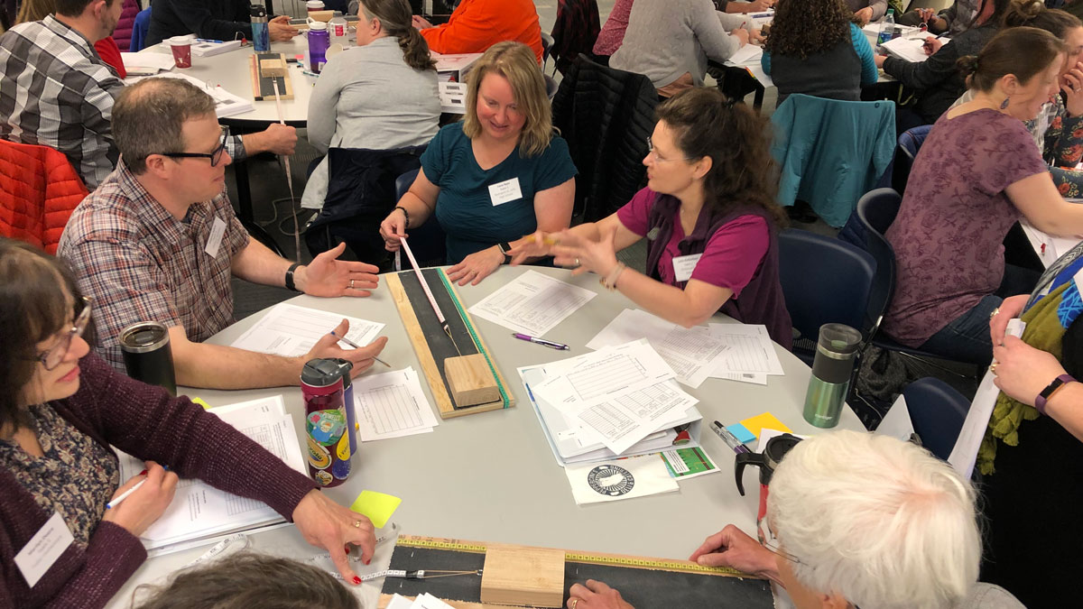 Leola Rutherford, dressed in a pink shirt and maroon vest, sits at a roundtable with ANGLE workshop attendees. Two participants are operating earthquake machines.
