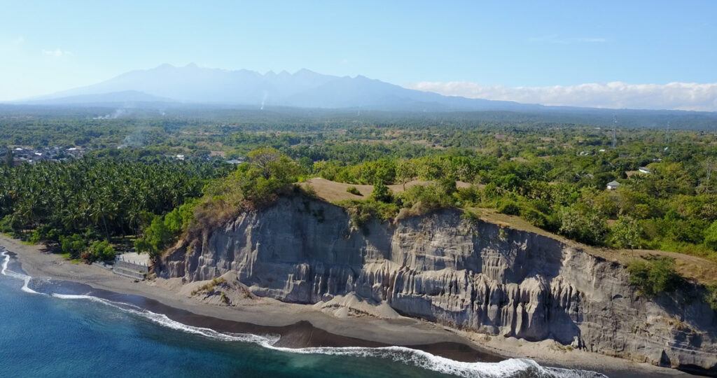 Cliffs of pumice tower about 35 meters high dominate an aerial view of the island of Samalas, while the Rinjani Volcanic Complex can be seen rising in the background.