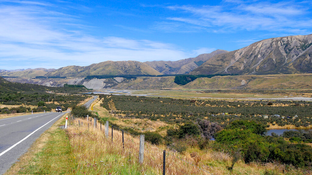Mountains and blue sky in the background, with green pastures in the foreground. A road extends from the foreground toward the mountains.