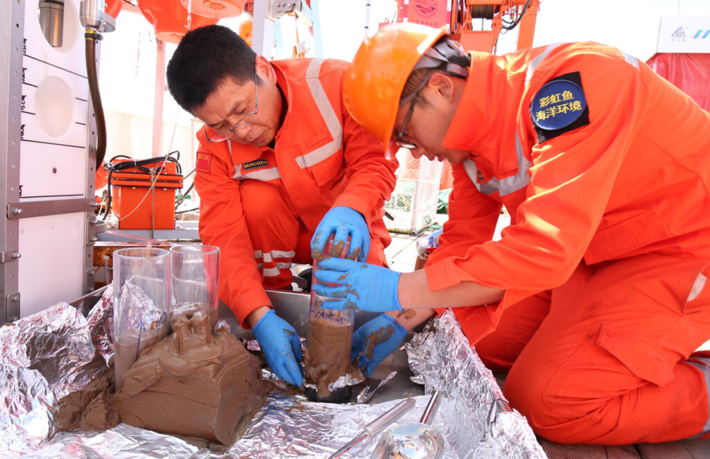 People in orange work suits crouch next to small clear cylinders with brown material inside.