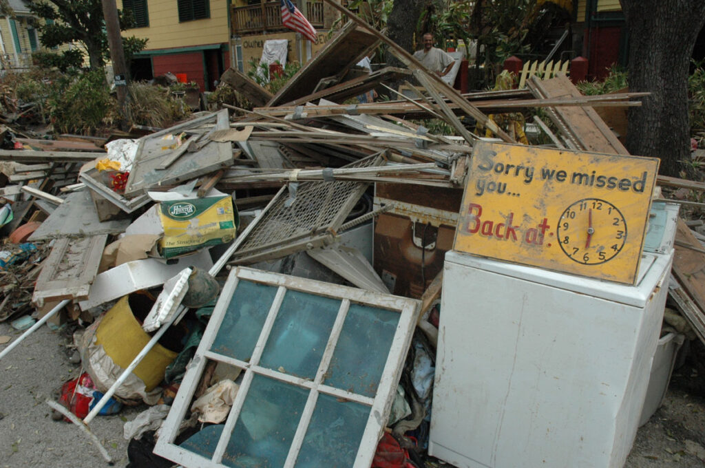 A large pile of debris, including window frames, wood slats, and more, sits in front of houses.