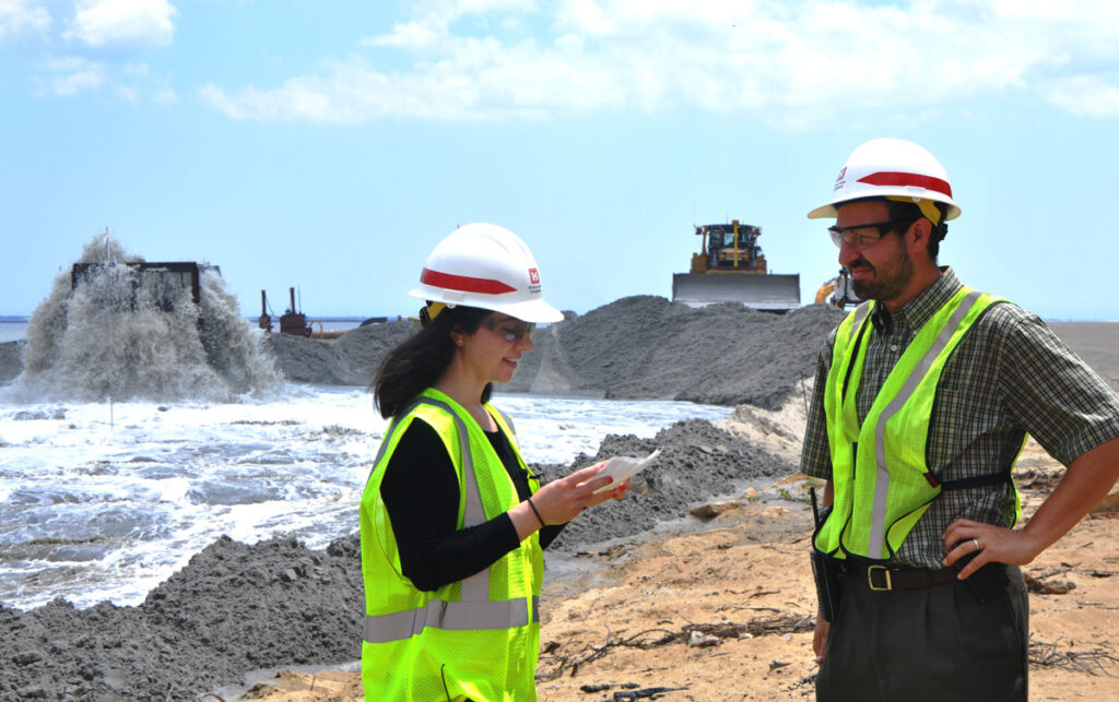 Two people wearing hard hats and yellow safety vests confer while standing on a beach in front of piles of sand and a bulldozer in the background.