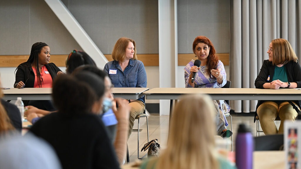 Four people, one speaking into a microphone, sit at a long table in front of audience members.
