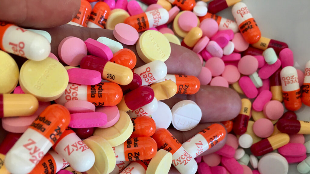 A person’s hand holding many pills and a pile of pills on the surface underneath