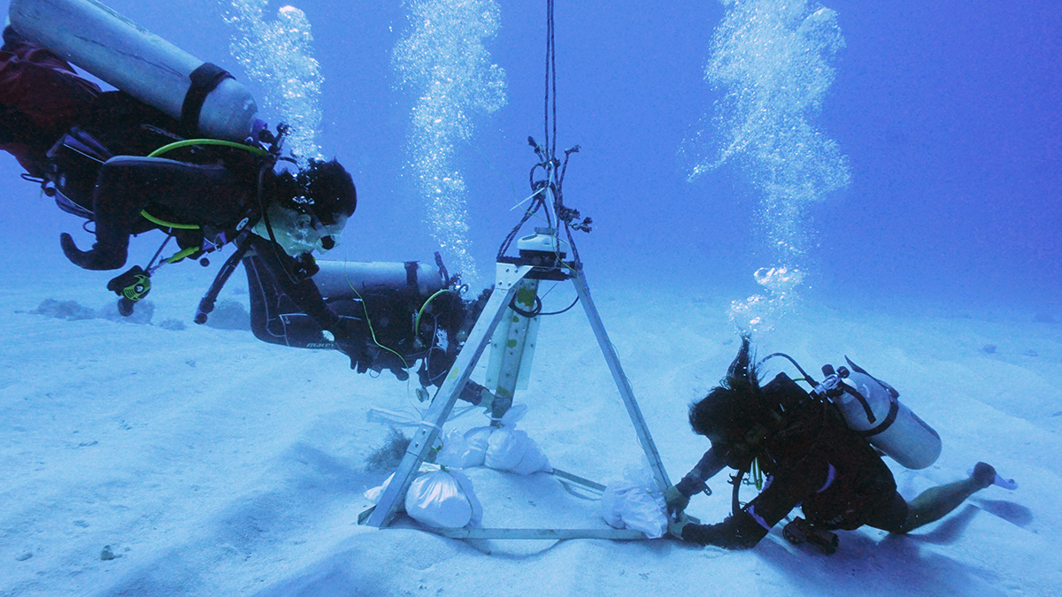 Underwater photo of metal equipment sitting on the seafloor with three divers around it.