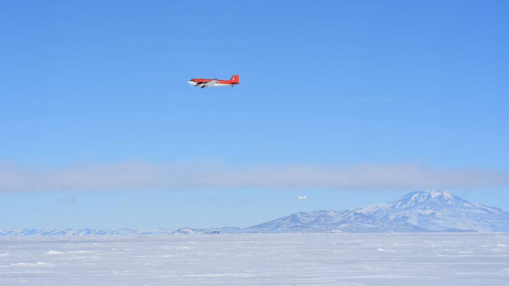 A red plane flies against clear blue skies over white ice. Mountains can be seen in the background.