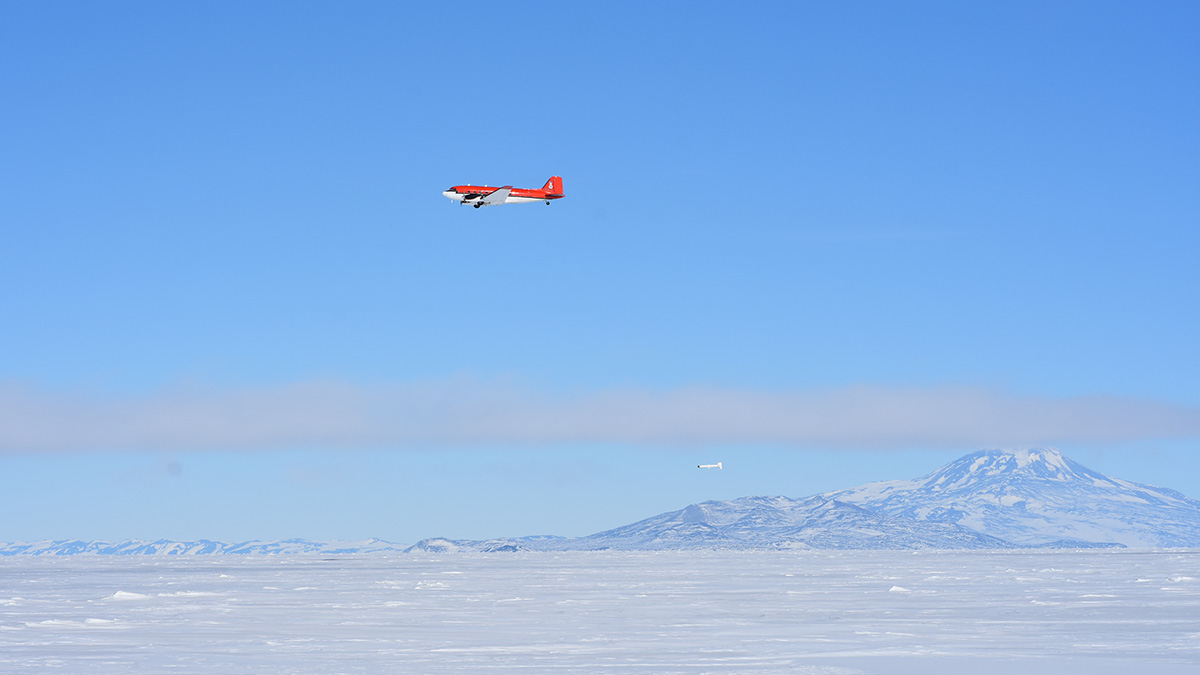 A red plane flies against clear blue skies over white ice. Mountains can be seen in the background.