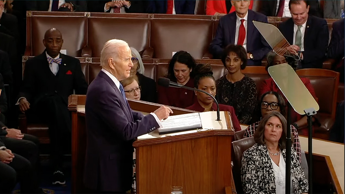 President Biden stands at a podium to deliver his speech to a crowded room of legislators and guests.