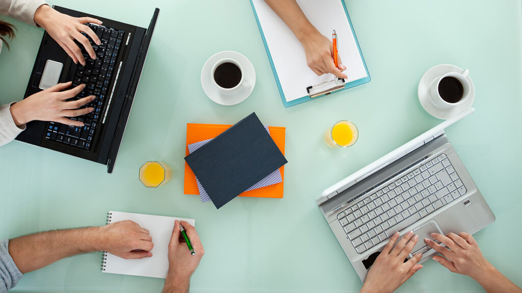 Overhead view of four people working together at a table with pads of paper, laptops, and coffee mugs, cropped to show only the hands and forearms of the people