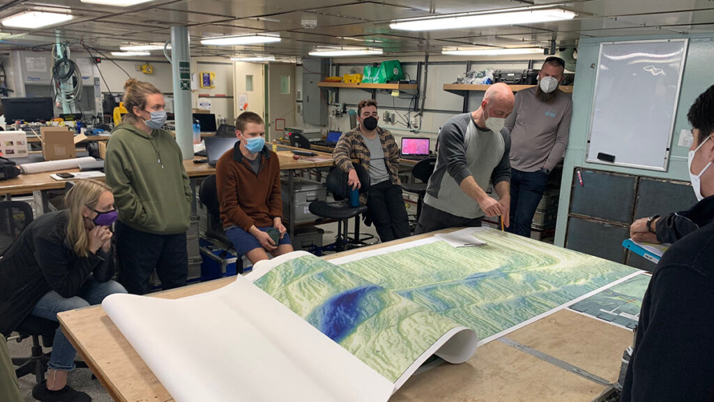 Several people sit and stand around a large map of the seafloor on a table in laboratory space.