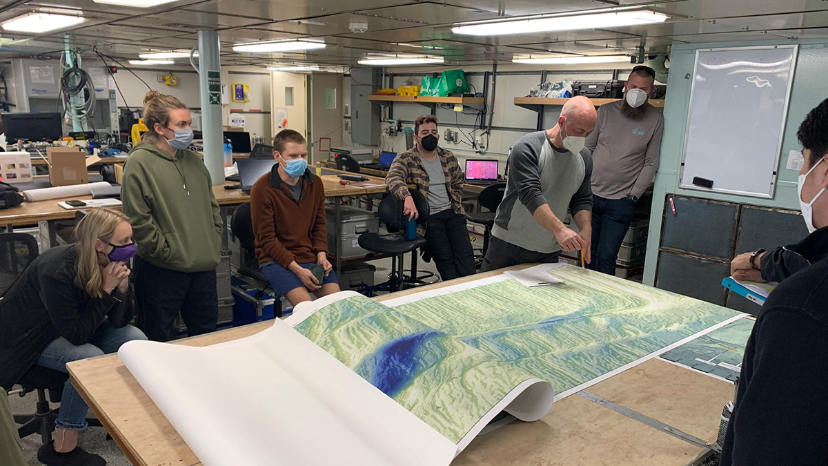 Several people sit and stand around a large map of the seafloor on a table in laboratory space.