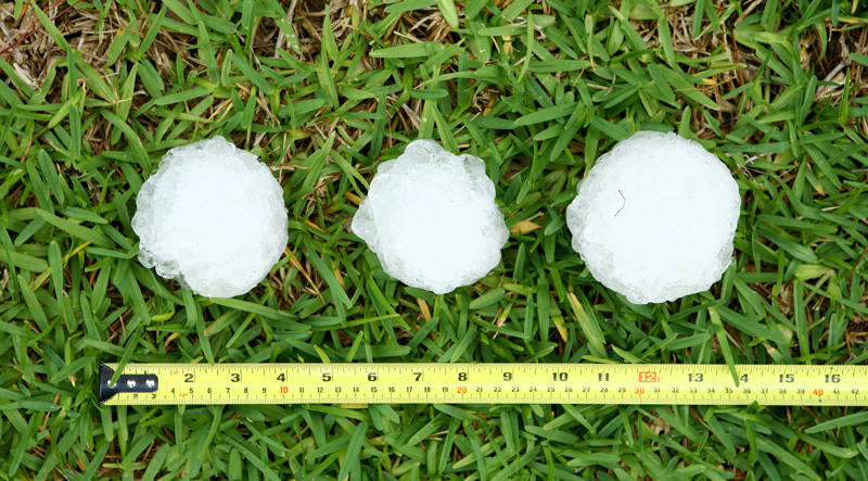 Three hailstones, each about the size of a baseball, sit atop grass with a measuring tape extended beside them to indicate their size.