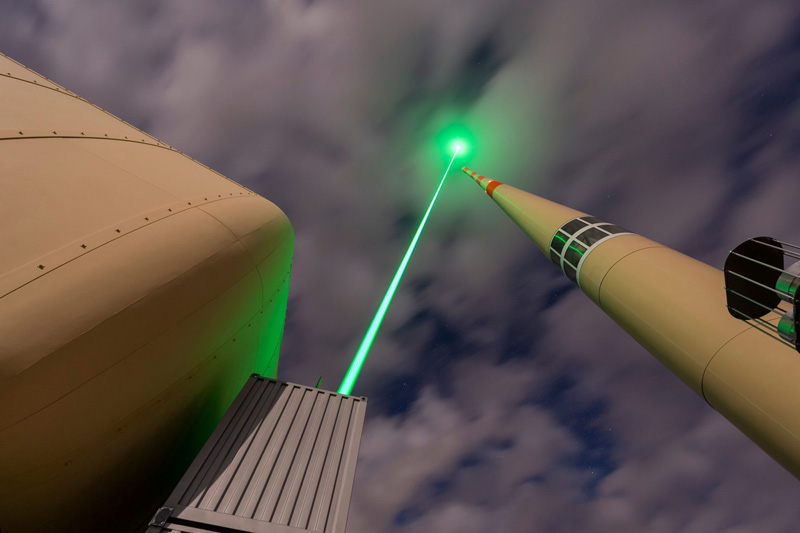 A bottom-up view of a large laser device firing a green laser into a cloudy sky